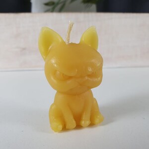 May include: A small, yellow, wax candle shaped like a sitting dog. The dog has a grumpy expression.