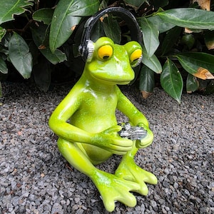 Op de afbeelding: Een groene kikkerstandbeeld met gele ogen en een headset. De kikker houdt een zilveren gamecontroller vast. Het beeld staat op een bedje van kleine grijze steentjes, met groene bladeren op de achtergrond.
