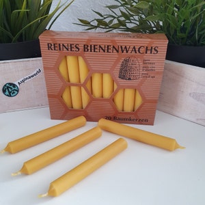 Op de afbeelding: Een doos met 20 bijenwaskaarsen, met het label "Reines Bienenwachs" en de tekst "pure beeswax, pure cire d'abeilles, pura cera d'api" en "20 Baumkerzen". De kaarsen zijn geel en zijn in een honingraatpatroon op de doos gerangschikt.