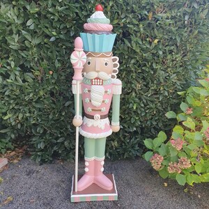 Pode incluir: Uma grande e extravagante estátua de quebra-nozes com um chapéu em forma de cupcake. A estátua é pintada em rosa pastel, verde e azul, com um bigode branco e um bastão de doces. Fica numa base castanha.