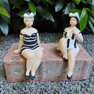 Può includere: Due figurine in ceramica di donne che indossano costumi da bagno a righe bianche e nere e fasce per capelli bianche. Sono sedute su un bordo di mattoni.