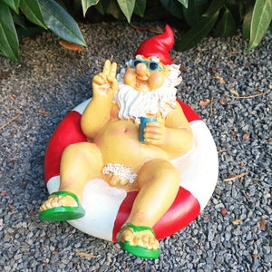 Puede incluir: Una caprichosa figura de gnomo de jardín, tomando el sol en un flotador inflable rojo y blanco. El gnomo lleva gafas de sol, un gorro rojo y sandalias verdes, sosteniendo una bebida azul. El gnomo hace el signo de la paz.