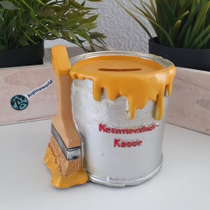 Könnte beinhalten: Eine gelbe Farbeimer-Sparbüchse mit einem Pinsel an der Seite. Der Eimer hat den Schriftzug "Heimwerker Kasse" aufgedruckt.