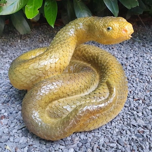 Könnte beinhalten: Eine gewundene, gelb-braune Schlangenskulptur mit detaillierter Schuppentextur. Der Kopf der Schlange ist erhoben, mit gelbem Maul und schwarzer Zunge. Die Statue steht auf einem Bett aus grauem Kies, mit grünem Laub im Hintergrund.