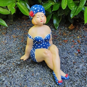 May include: A ceramic figurine of a woman in a blue swimsuit with white heart patterns, a matching hat, and blue shoes. The hat has a red rose. The figurine is sitting on a bed of small gray stones.