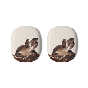 May include: Two oval-shaped decorative items featuring a fawn peeking out. The fawn has brown fur with white spots and large ears. The background is a light beige color.