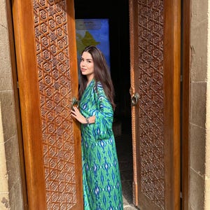 Puede incluir: Un vestido largo y fluido de color verde con un estampado azul y blanco. El vestido tiene mangas largas y se lleva delante de una puerta de madera con intrincadas tallas. La puerta est&aacute; abierta, revelando un vistazo a un mapa.