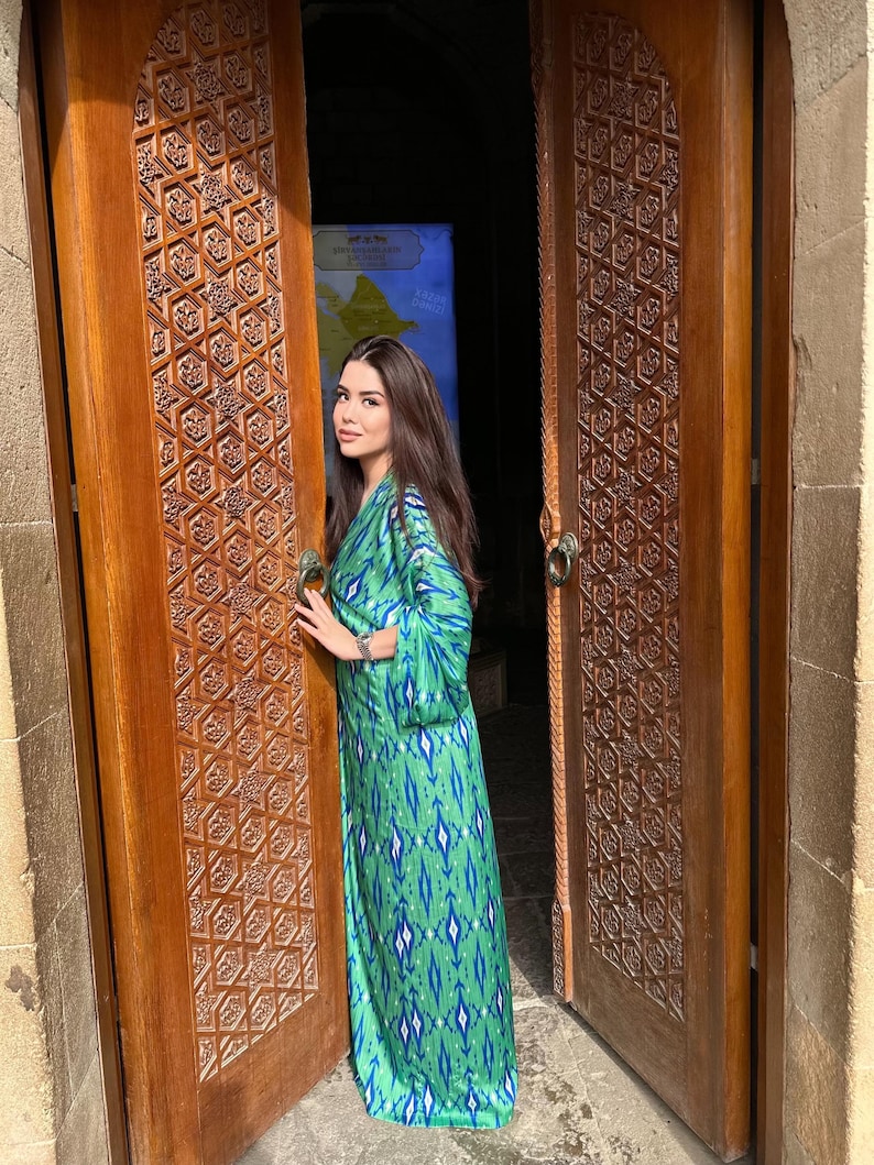 Puede incluir: Un vestido largo y fluido de color verde con un estampado azul y blanco. El vestido tiene mangas largas y se lleva delante de una puerta de madera con intrincadas tallas. La puerta est&aacute; abierta, revelando un vistazo a un mapa.