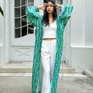 Puede incluir: Una bata larga y fluida de estilo kimono con estampado verde y azul. La bata se lleva sobre un top corto blanco y pantalones blancos. Un adorno dorado para la cabeza y sandalias completan el look. El fondo es un edificio blanco con una puerta.