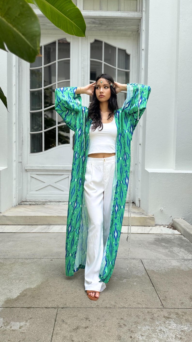 Puede incluir: Una bata larga y fluida de estilo kimono con estampado verde y azul. La bata se lleva sobre un top corto blanco y pantalones blancos. Un adorno dorado para la cabeza y sandalias completan el look. El fondo es un edificio blanco con una puerta.
