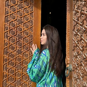 Puede incluir: Una persona con un kimono verde y azul estampado est&aacute; de pie cerca de una puerta de madera tallada. La puerta tiene intrincados dise&ntilde;os geom&eacute;tricos. El patr&oacute;n del kimono es una mezcla de azul y blanco sobre un fondo verde. La puerta est&aacute; abierta, revelando un fondo oscuro.