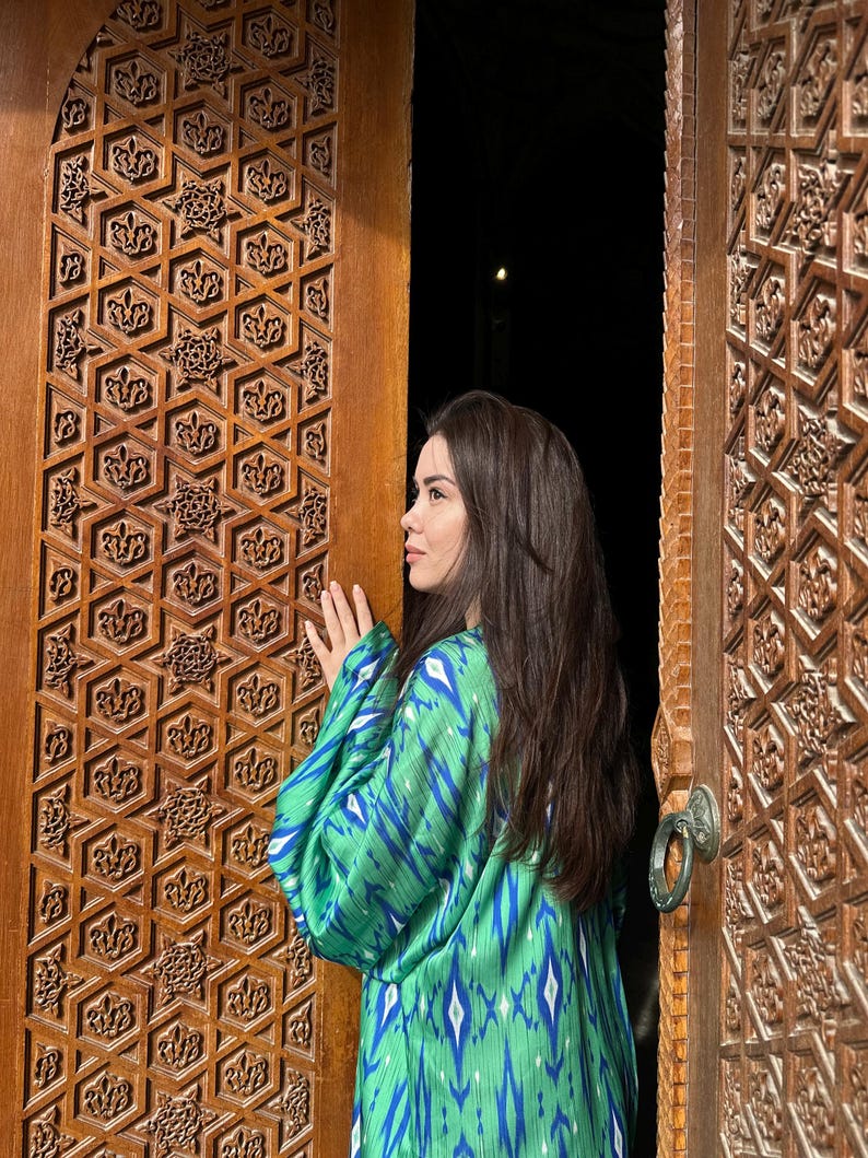 Puede incluir: Una persona con un kimono verde y azul estampado est&aacute; de pie cerca de una puerta de madera tallada. La puerta tiene intrincados dise&ntilde;os geom&eacute;tricos. El patr&oacute;n del kimono es una mezcla de azul y blanco sobre un fondo verde. La puerta est&aacute; abierta, revelando un fondo oscuro.