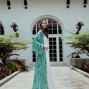 Puede incluir: Una larga bata estilo kimono verde y azul estampada sobre un atuendo blanco. La bata presenta un dise&ntilde;o geom&eacute;trico. La persona est&aacute; de pie frente a un edificio blanco con grandes ventanas y puertas arqueadas.