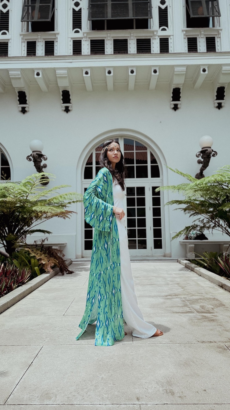 Puede incluir: Una larga bata estilo kimono verde y azul estampada sobre un atuendo blanco. La bata presenta un dise&ntilde;o geom&eacute;trico. La persona est&aacute; de pie frente a un edificio blanco con grandes ventanas y puertas arqueadas.