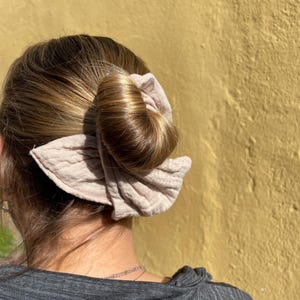 Op de afbeelding: Een lichtbruine scrunchie-knot met een textuurontwerp wordt in het haar gedragen. De scrunchie heeft een neutrale kleur en lijkt gemaakt te zijn van een zachte stof. Het haar is lichtbruin en opgestoken in een knot.