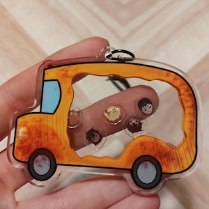 Puede incluir: Un llavero de autobús escolar amarillo con una ventana transparente que muestra personajes de dibujos animados en el interior. El autobús tiene contornos negros y un parabrisas azul.