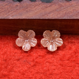 May include: Two pink flower-shaped cabochons with a sparkly finish. The cabochons are displayed on a red velvet surface.