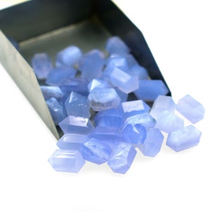 Puede incluir: Una pala llena de piedras preciosas de calcedonia azul claro, que se derraman sobre una superficie blanca. Las piedras preciosas son facetadas y tienen una apariencia translúcida.