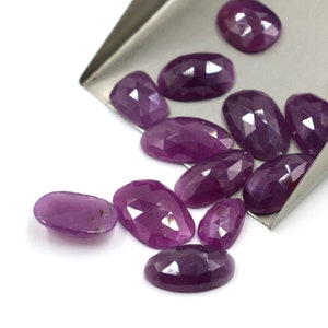 May include: A group of purple faceted oval gemstones, some are in a metal scoop and others are scattered on a white surface.