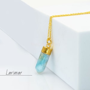 Puede incluir: Un collar de cadena dorada con un colgante de piedra Larimar azul claro. El colgante es un cristal largo, delgado y facetado con una tapa chapada en oro.