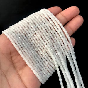 Puede incluir: Múltiples hilos de pequeñas cuentas de piedra lunar blanca translúcida de forma cuadrada. Cada hilo presenta cuentas uniformes y pulidas con un sutil brillo iridiscente, dispuestas en líneas paralelas para mostrar la textura suave y la calidad reflectante de la luz del material de piedra preciosa.
