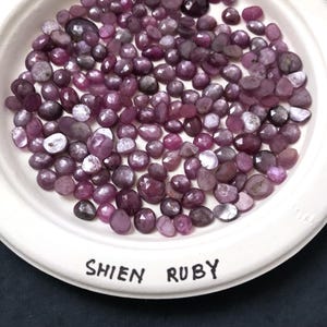 May include: A collection of faceted, deep red-purple Shien Ruby gemstones displayed on a white plate. The gemstones vary in size and shape, with a mix of light and dark hues. The plate has the words "Shien Ruby" printed on it.