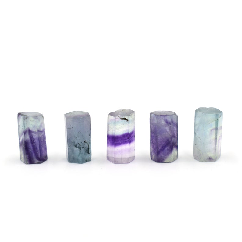 Puede incluir: Cinco puntas de cristal de fluorita en varios tonos de morado, azul y blanco. Los cristales est&aacute;n dispuestos en una fila sobre un fondo blanco.