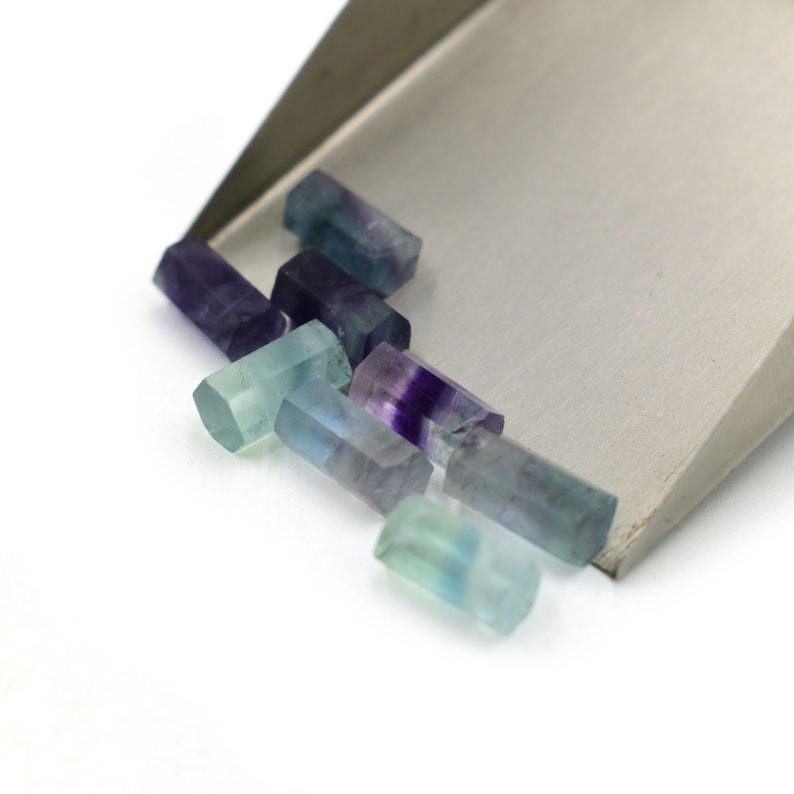 Puede incluir: Un grupo de peque&ntilde;os cristales de fluorita rectangulares y facetados en tonos de verde, azul y morado. Los cristales est&aacute;n dispuestos en una pila sobre una superficie de metal plateado.