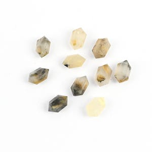 May include: A collection of ten small, hexagonal crystals. The crystals have a translucent appearance with a mix of pale yellow, gray, and brown inclusions. They are scattered on a white surface, showcasing their natural variations.