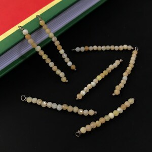 May include: Several strands of faceted, light yellow-beige beads with silver-colored loops at the ends. The beads are scattered on a black surface. A book with a green, yellow, and red cover is in the background.