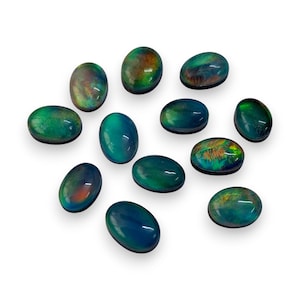 Puede incluir: Un conjunto de ópalos ovalados en tonos verdes, azules y toques de naranja y rojo. Las piedras preciosas están dispuestas sobre una superficie blanca, mostrando sus cualidades iridiscentes. Estos ópalos son adecuados para la creación de joyas.
