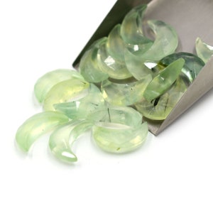 May include: A collection of green crescent moon shaped stones with black inclusions. The stones are arranged in a pile and are spilling out of a metal scoop.