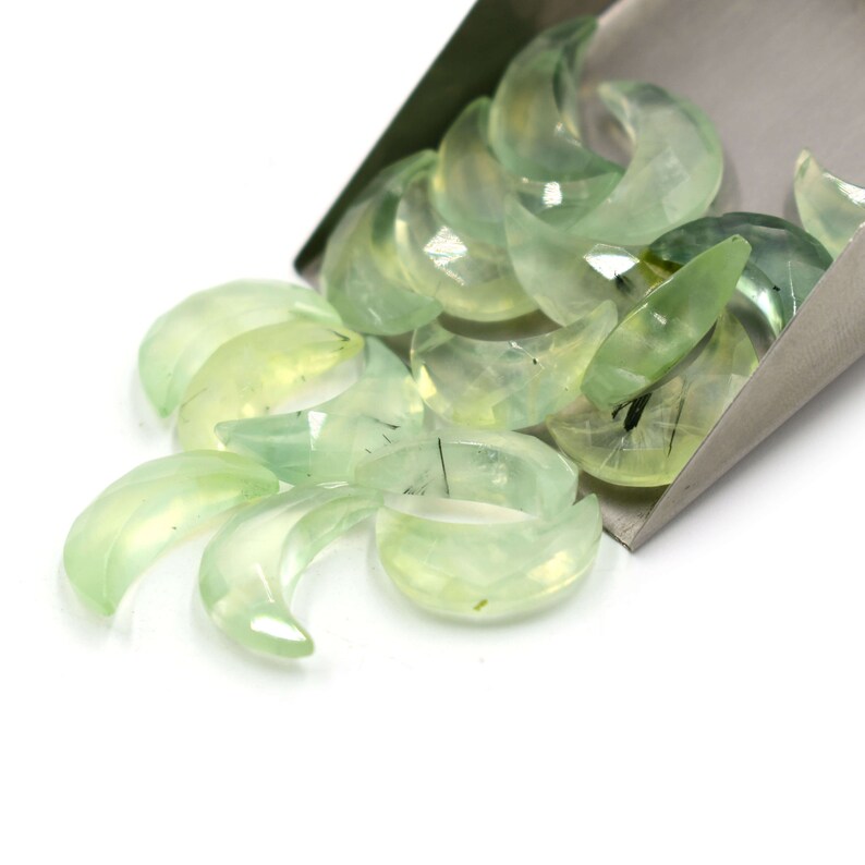 May include: A collection of green crescent moon shaped stones with black inclusions. The stones are arranged in a pile and are spilling out of a metal scoop.