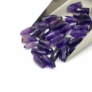 Könnte beinhalten: Eine Schaufel mit Amethyst-Edelstein-Spitzen, jede mit einer spitzen Spitze und einem facettierten, violetten, durchscheinenden Körper. Die Edelsteine sind in einem lockeren Haufen angeordnet, der aus einer Metallschaufel herausfällt.