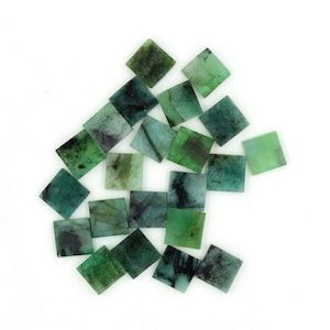May include: A collection of 18 square-shaped emerald green gemstones with black inclusions. The gemstones are arranged in a loose pile on a white background.