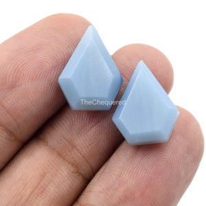 Può includere: Due cabochon di pietre preziose azzurre a forma di aquilone geometrico. Le pietre lucide hanno una lucentezza sottile. Il testo "TheChequered" è visibile. Questi cabochon sono adatti per la creazione di gioielli.