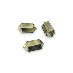 Puede incluir: Tres cristales de pirita dorados y facetados. Cada cristal tiene una forma rectangular con múltiples lados angulados, que reflejan la luz. Los cristales están dispuestos sobre un fondo blanco.