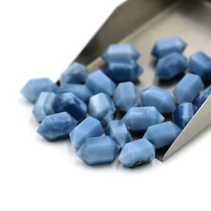 May include: A group of blue, faceted, gemstone beads in a metal scoop. The beads are all roughly the same size and shape, and they have a slightly iridescent sheen.