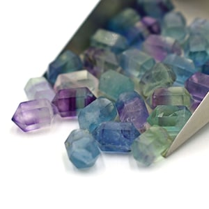 Könnte beinhalten: Eine Sammlung von facettierten, irisierenden Fluoritkristallen in Blautönen, Grüntönen und Violett. Die Kristalle sind in einem lockeren Haufen angeordnet und fallen aus einer Metallschaufel.