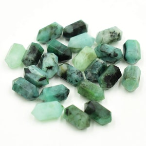 Puede incluir: Colección de cristales de piedras preciosas de color verde esmeralda con diferentes tonos e inclusiones oscuras. Los cristales tienen forma hexagonal y parecen pulidos. Las piedras preciosas están dispersas sobre una superficie blanca.