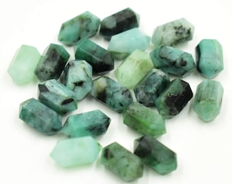 Puede incluir: Colección de cristales de piedras preciosas de color verde esmeralda con diferentes tonos e inclusiones oscuras. Los cristales tienen forma hexagonal y parecen pulidos. Las piedras preciosas están dispersas sobre una superficie blanca.
