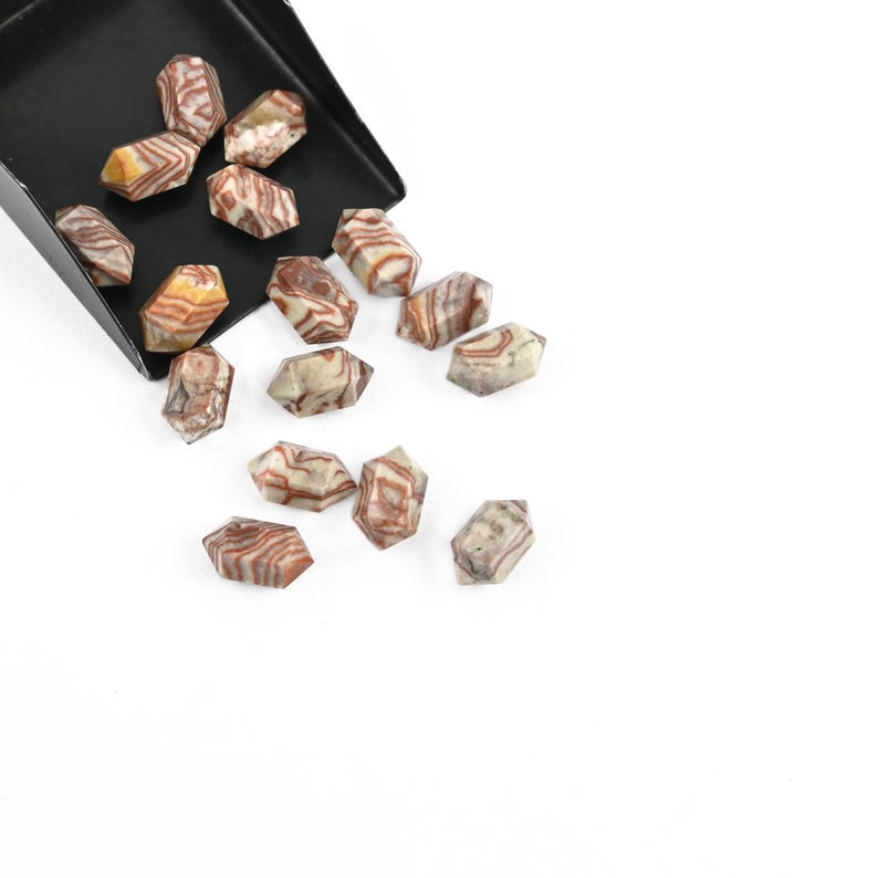 May include: Tumbled gemstones with a banded pattern in brown, cream, and tan hues. The hexagonal stones are spilling from a black scoop onto a white surface. Each stone has a unique pattern.