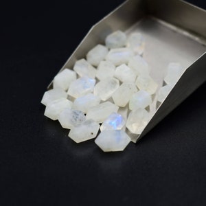 May include: A collection of small, faceted, white moonstone gemstones with a subtle iridescent sheen. The gemstones are in a metal scoop, with some spilling out onto a black surface. The stones have a hexagonal shape.