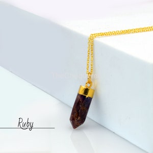 Może przedstawiać: Złoty łańcuszek z wisiorem w kształcie rubinowej pointy. Wisiorek jest ciemnoczerwony i ma pozłacaną końcówkę z obu stron. Pod wisiorem widnieje napis "Ruby".