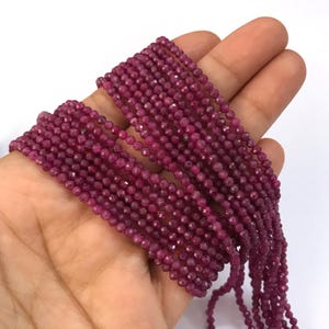 May include: A strand of small, round, faceted ruby-colored beads. The beads are deep magenta in color and have a reflective, multi-faceted surface. The strand is arranged in multiple parallel rows, showcasing the uniform size and texture of the beads.