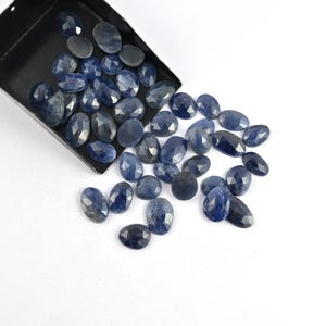May include: A collection of faceted, oval-shaped blue sapphires. The gemstones vary in size and shade, with a deep, rich blue hue. Some sapphires are spilling out of a black tray onto a white surface.