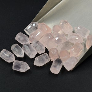 Può includere: Una collezione di cristalli di quarzo rosa pallido di forma allungata e sfaccettata. I cristalli sono sparsi su una superficie nera.