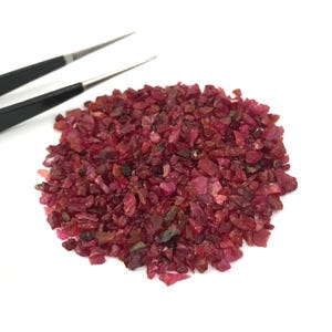 Peut inclure: Une collection de petits éclats de rubis rouge foncé, irréguliers, disposés en un tas circulaire. Les pierres présentent des nuances variées de cramoisi et de bordeaux avec une texture translucide et facettée. Une pince en métal noir est visible.
