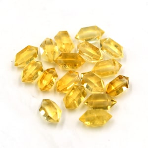 May include: A group of 16 faceted, yellow citrine crystals in a double terminated, hexagonal shape. The crystals are arranged in a loose pile on a white background.