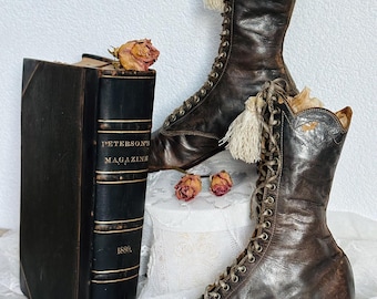 May include: A pair of brown leather lace-up boots with white tassels. The boots have a high heel and are sitting on a white lace tablecloth. A vintage book titled "Peterson's Magazine 1880" is in the background.